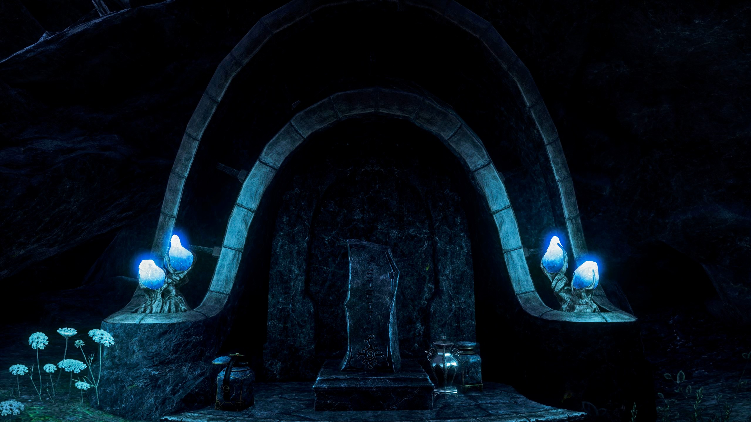 A screenshot from the game Elder Scrolls Online. It depicts the entrance of an Ancestor Tomb, located in Vvardenfell. The entrance is carved into the surrounding rock. It consists of a stone arch which surrounds a stone door. The door is shaped like the arch. There is a stone slab in front of the door, which is shaped like a rectangle, but with jagged edges. Left and right of the door are small planters, which are extensions of the stone arch. They contain blue, fluorescent fungi. The atmosphere is calm and a little bit eerie.